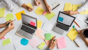 Equipe diversa discutindo ideias em uma mesa com laptops e blocos de notas, com quadro branco cheio de diagramas e post-its ao fundo.